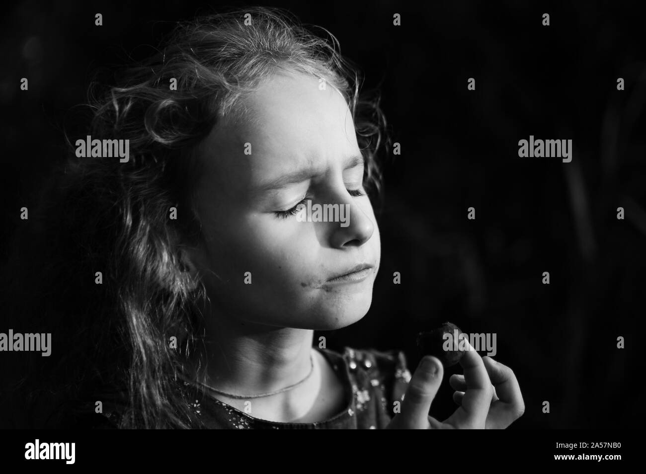 White and black portrait of little blonde girl with ponytail eating a chocolate candy with closed eyes outdoors, happy childhood concept Stock Photo