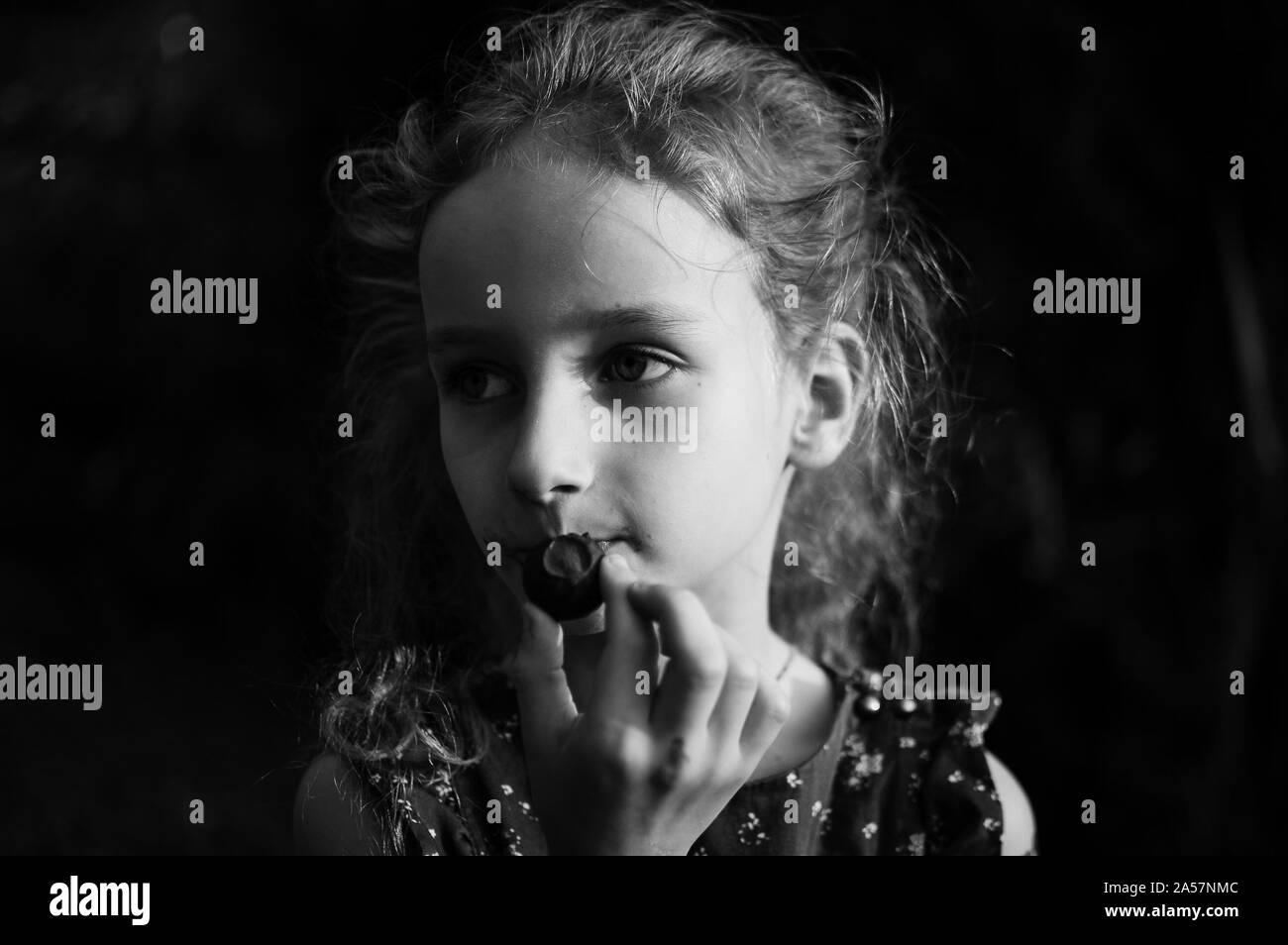 Close up black and white portrait of little blonde girl with ponytail eating a chocolate candy outdoors, sugar addiction concept Stock Photo