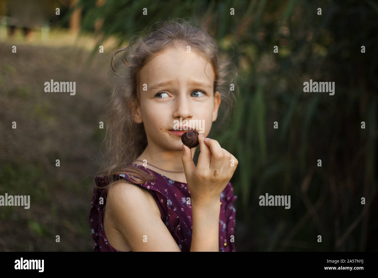 Beautiful little blond girl in violet dress with ponytail is eating a chocolate candy in the park durring a day, sugar addiction concept Stock Photo