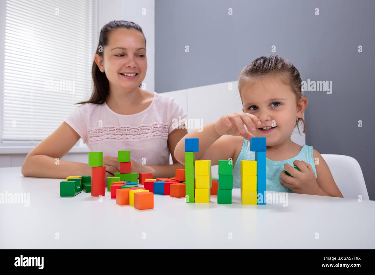 Smiling Daycare Worker Playing With Child Stacking Building Blocks On White Desk Stock Photo