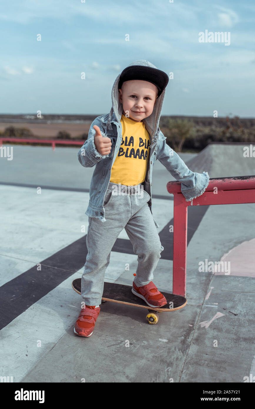 little boy of 4-5 years old, in autumn summer in city on sports ground,  learns to ride skateboard, smiles happy, shows finger to top. Casual wear  Stock Photo - Alamy