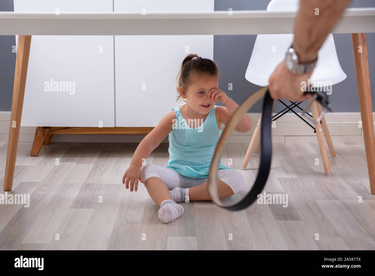 Father's Hand Holding Leather Belt In Front Of His Crying Daughter Sitting Under The Table Stock Photo