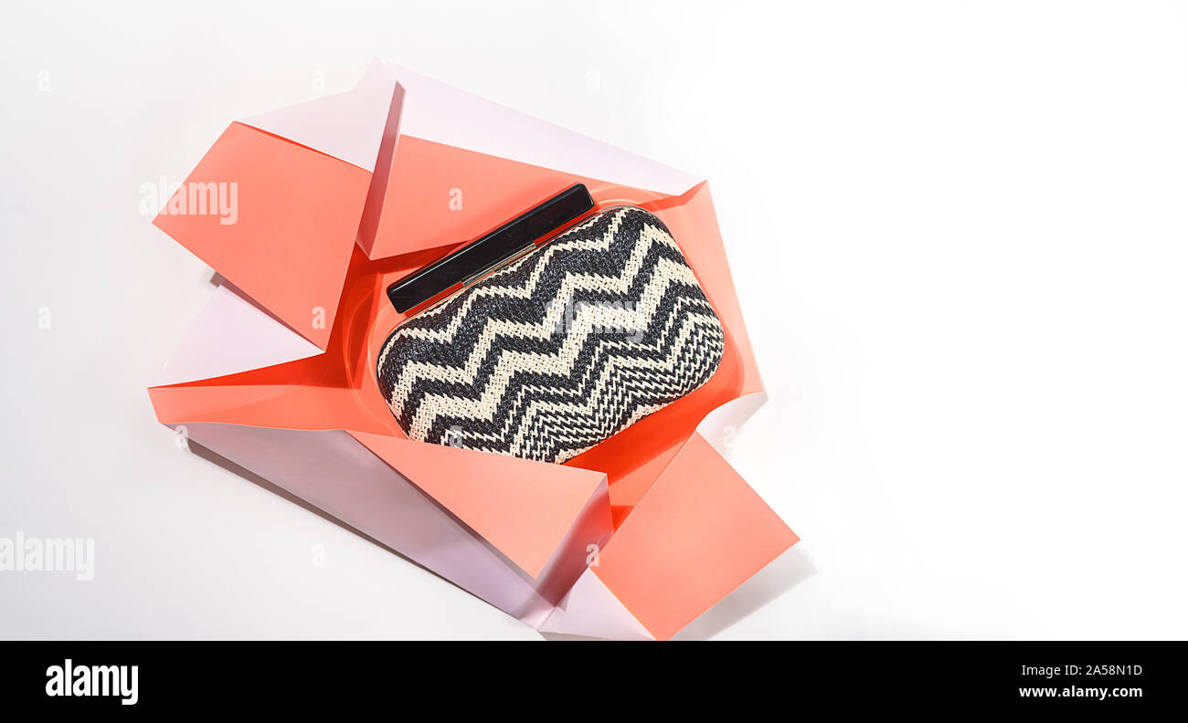 The concept of gift unwrapped from paper of living coral color on white background. Present for birthday, Valentine's Day, holiday. Black and white handbag or clutch bag. Top view, creative layout. Stock Photo