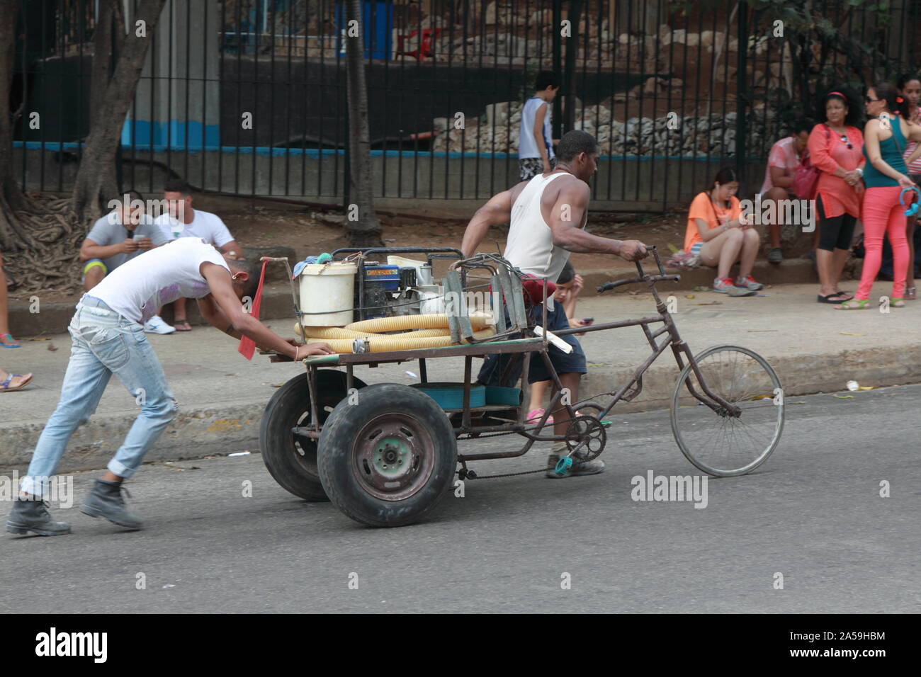 Two men in white T-shirts push their three-wheeled transport bike up a steep hill in central Havana. Stock Photo