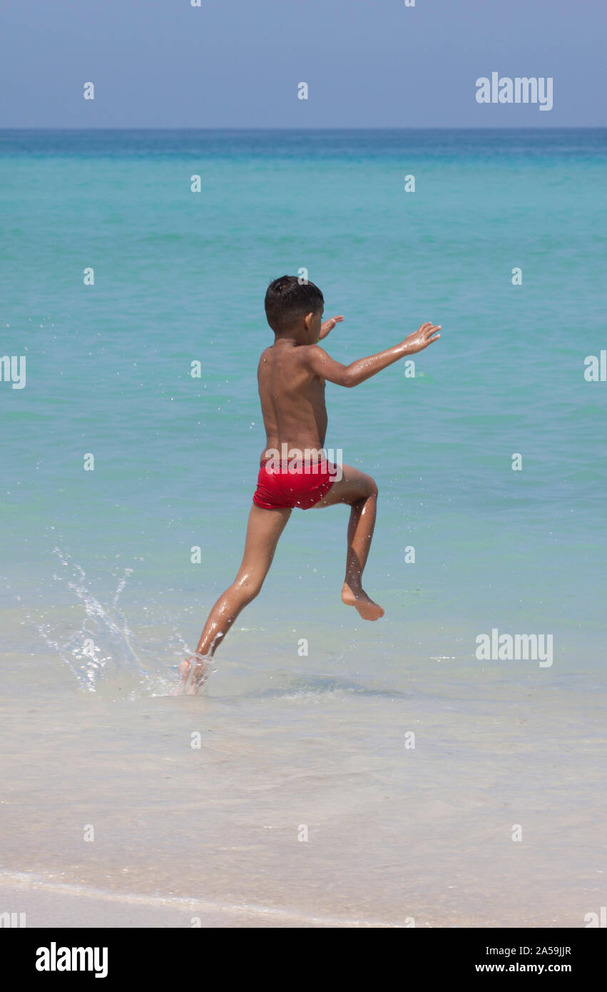 Young boy in red swimming trunks running out into the blue sea. Stock Photo