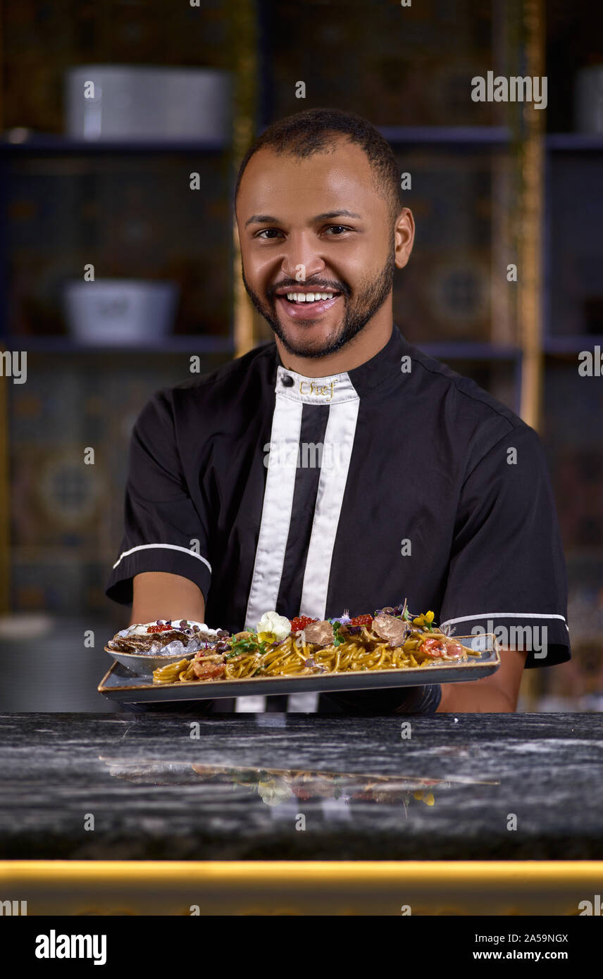 happy african american chef looking at camera, presenting Spaghetti, model in chef uniform holding a dish of seafood spaghetti, the dish in commercial Stock Photo