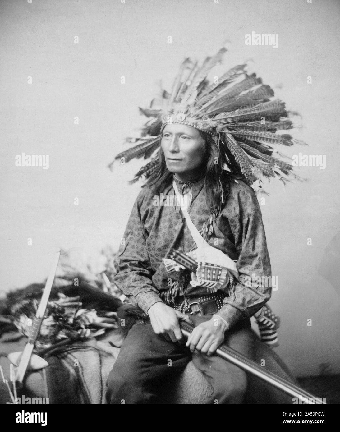 Little, Oglala band leader, three-quarter length studio portrait, seated, facing front, wearing turkey feather headdress, pants and shirt, holding rifle across lap. Stock Photo