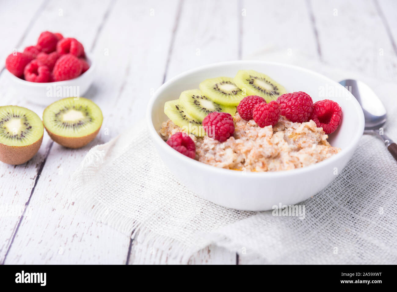 Organic healthy oatmeal porridge with fresh raspberries and kiwi. The white porcelain bowl is on a white wooden table, with a textile cloth underneath Stock Photo