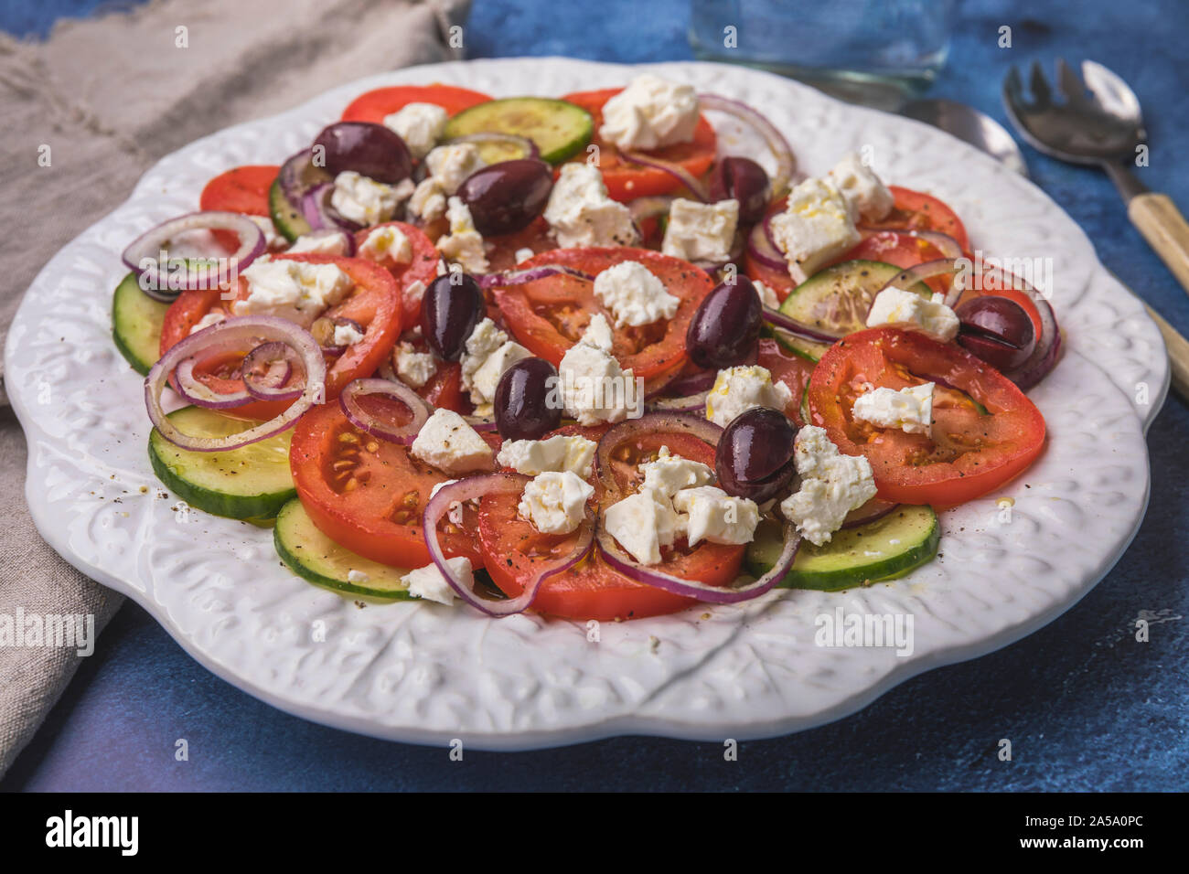 A fresh healthy salad with tomatoes, cucumber, red onion, feta cheese and kalamata olives. The salad is on a beautiful white plate on a blue backgroun Stock Photo