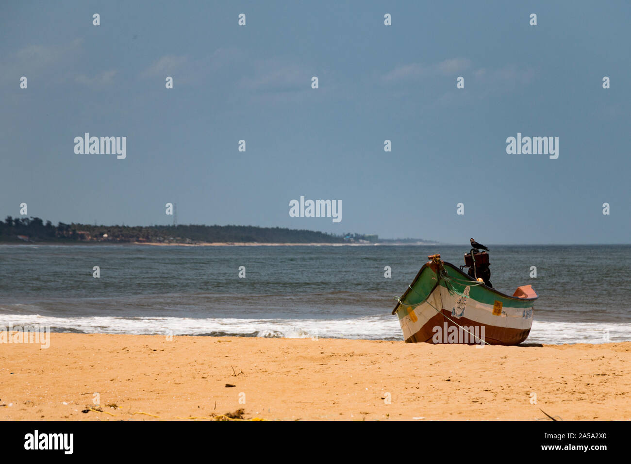 green, white and red fishing boat on a sandy beach on a sunny day with a crow sitting on the engine - India, Mamallapuram September 2019 Stock Photo