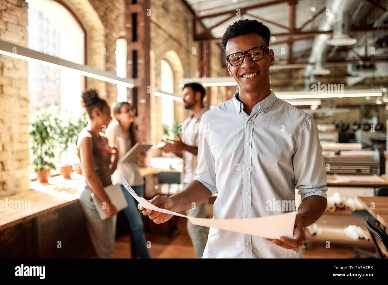 Enjoying my work. Young afro american man holding documents and looking at camera with smile while standing in the modern office Stock Photo