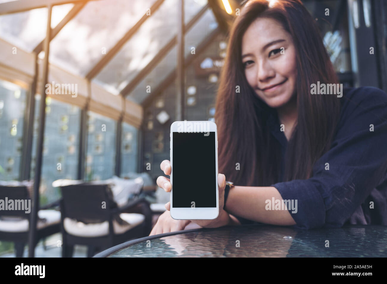 Mockup image of a beautiful asian woman holding and showing white mobile phone with blank black screen in cafe Stock Photo