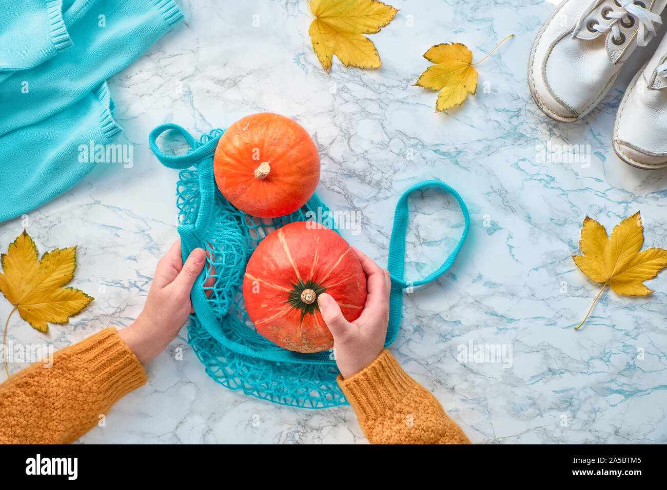 Autumn flat lay with female hands putting orange pumpkin into turquoise mesh bag. Top view on light marble background with white shoes, sweater and ye Stock Photo