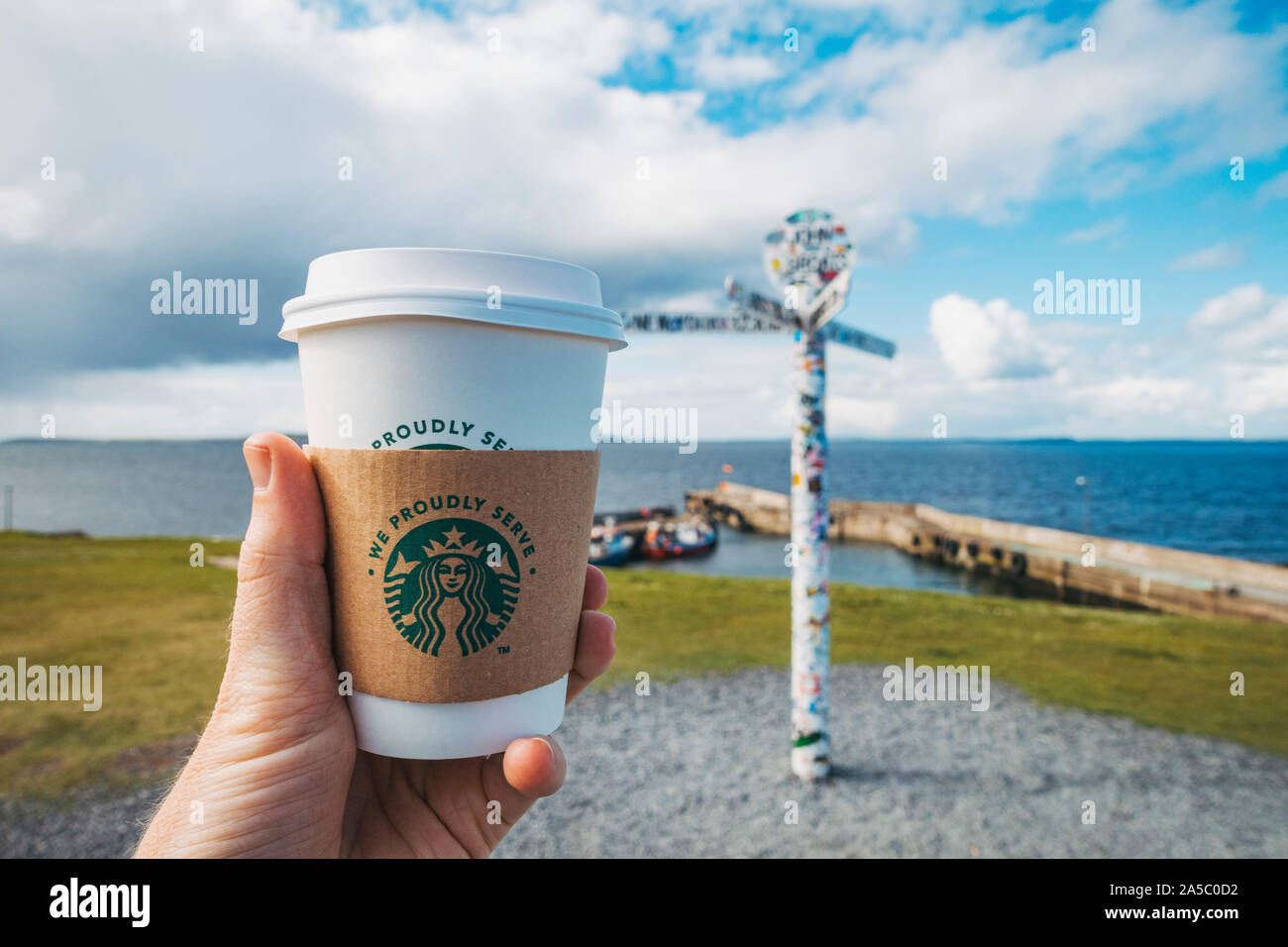 A tourist holds up a 'We Proudly Serve Starbucks' cup in front of the famous John o'Groats sign, in the far north of the United Kingdom Stock Photo
