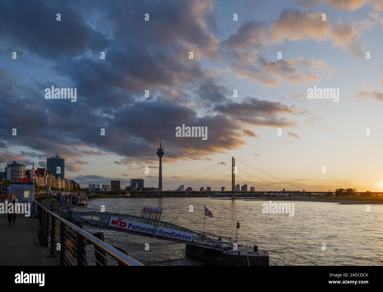 View of promenade on riverside, pedestrian bridge to pier of KD, Tourism company for Cruise on Rhine River, and background of Rhine Tower. Stock Photo