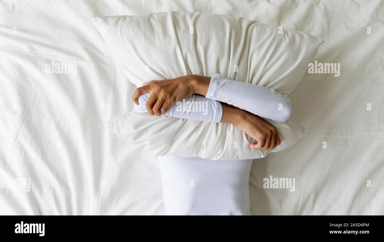 Young girl lying on bed, covering face with pillow. Stock Photo