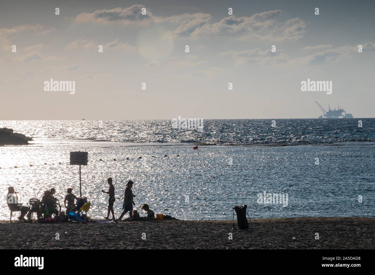 DOR BEACH, ISRAEL / 18 OCT 2019: Beachgoers relaxing on beach in front of environmentally controversial offshore drilling platform in the background. Stock Photo