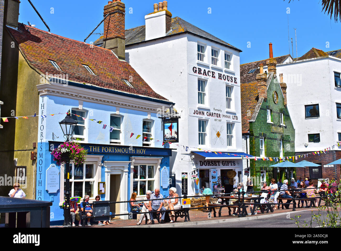 A pretty view of the colourful pubs and other buildings lined up along the quayside overlooking the harbour. People walking past or shopping. Stock Photo