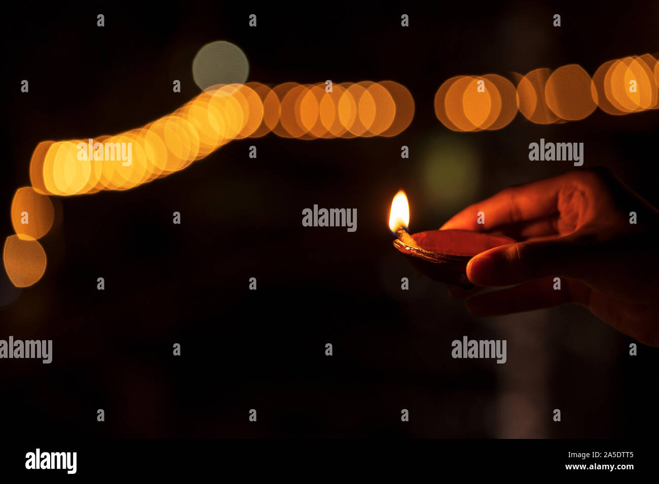 Diwali - the festival of lights. Person holding diya or clay oil lamp with yellow orange bokeh & space for text isolated in black background. Stock Photo