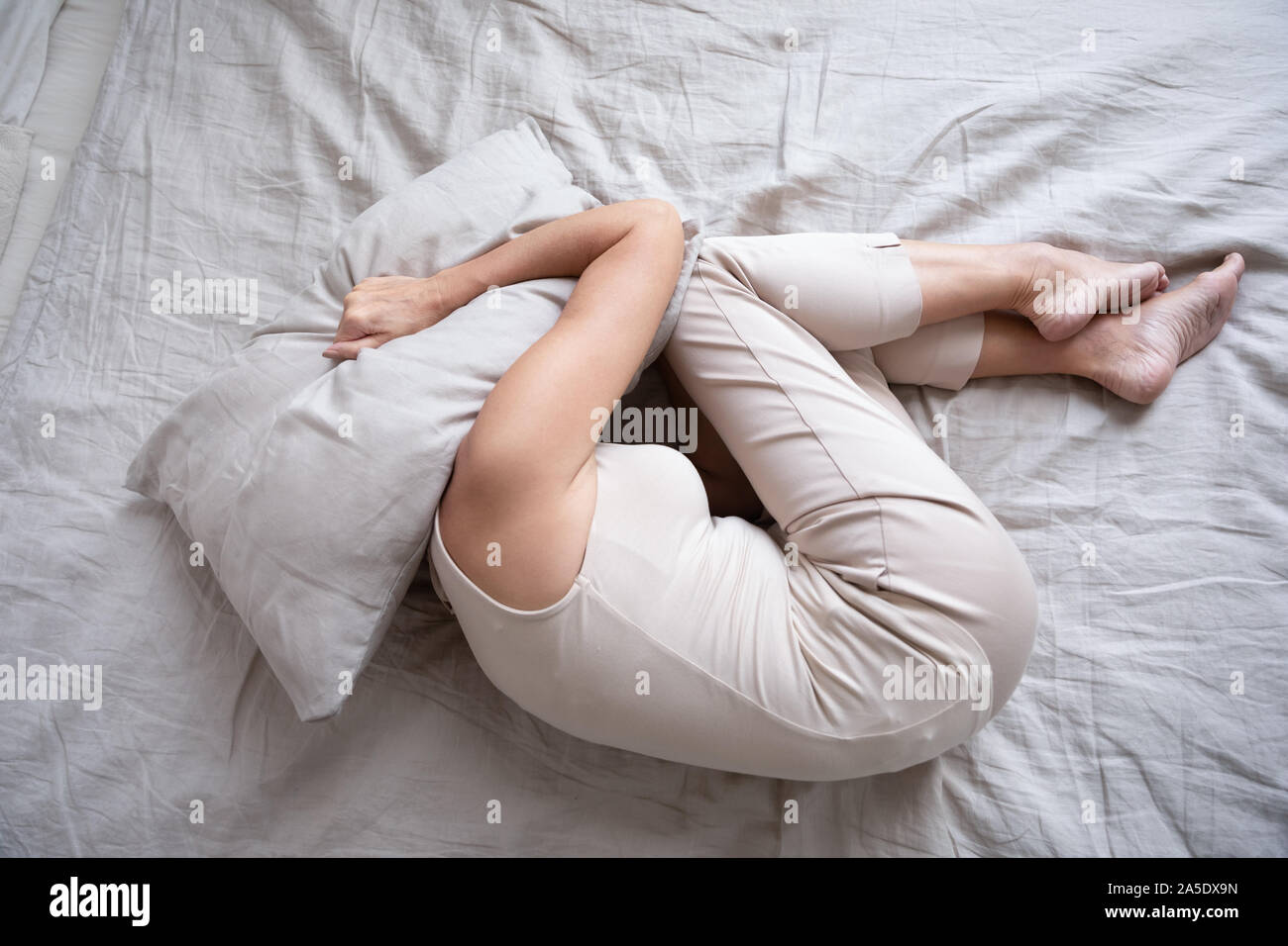Frightened mature woman lying on bed fetal position covering pillow Stock Photo