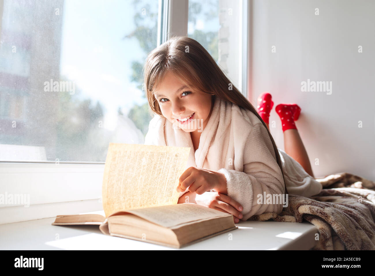 Cute little girl lying on the window and reading a book in room at home ...