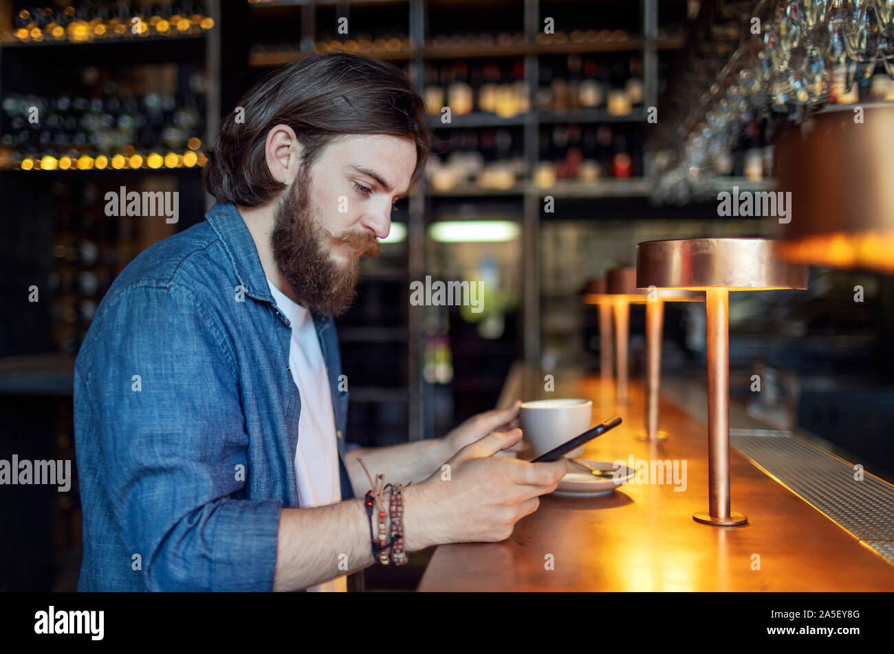Young brown-haired bearded man having his coffee and using smartphone Stock Photo