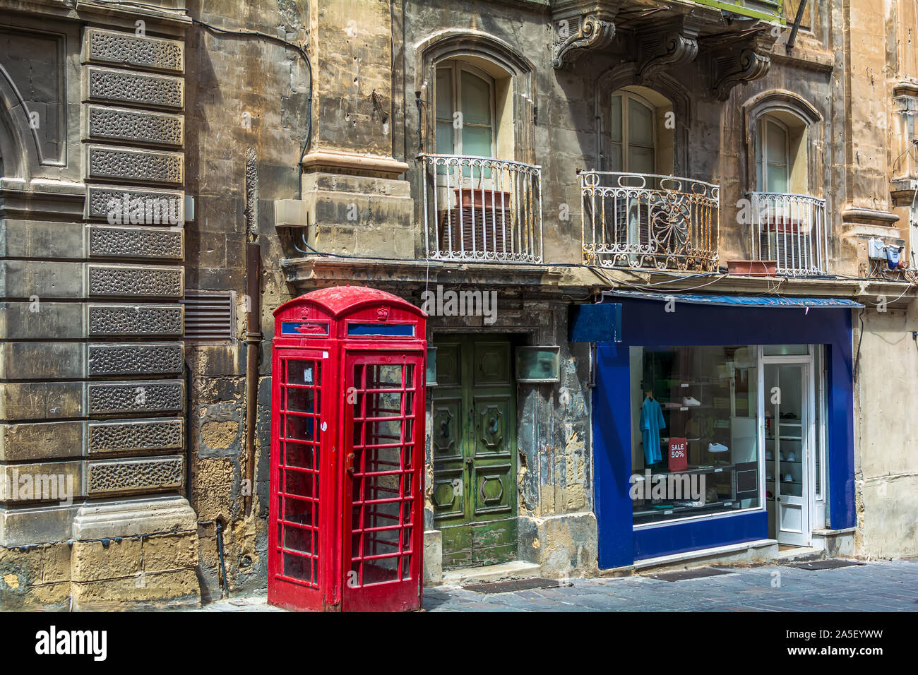London-style iconic red phone box standing next to run-down old building and bright show-window of clothing store in Valletta, Malta. Stock Photo