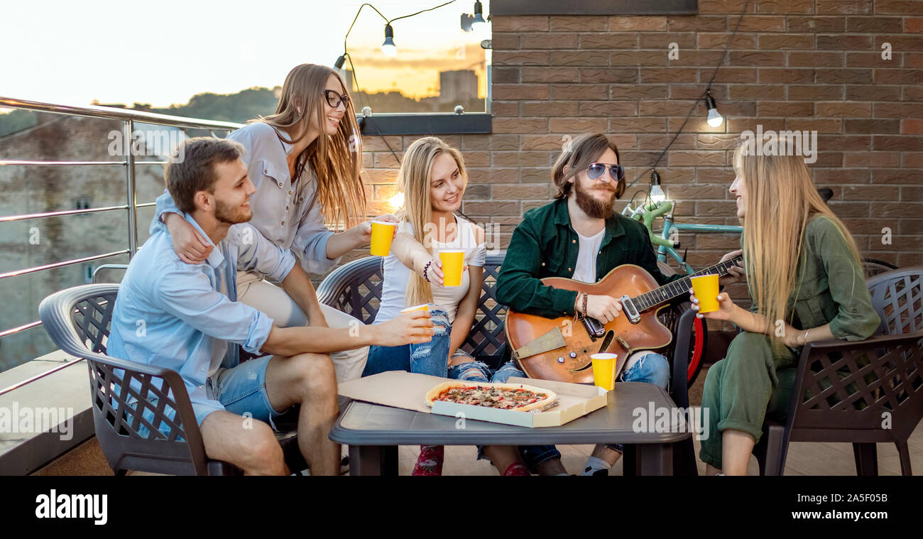 True friends having beer and pizza and listen their pal playing the guitar on a sunset loft terrace Stock Photo