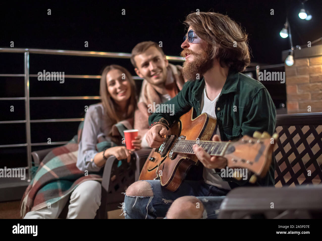 Young couple listening to their friend's romantic guitar-accompanied song on a cozy balcony after sunset Stock Photo