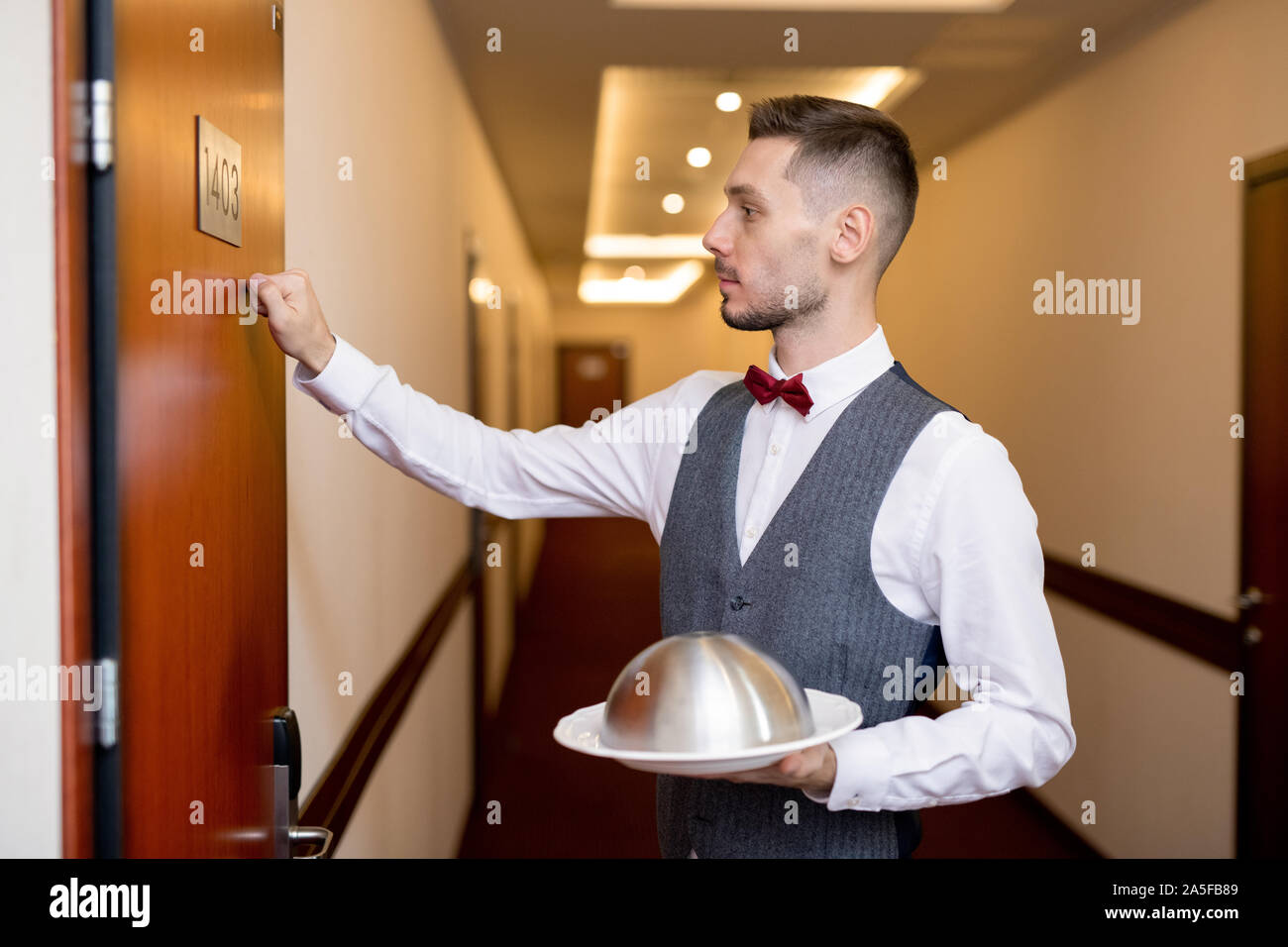 Young elegant waiter knocking on wooden door of hotel room while