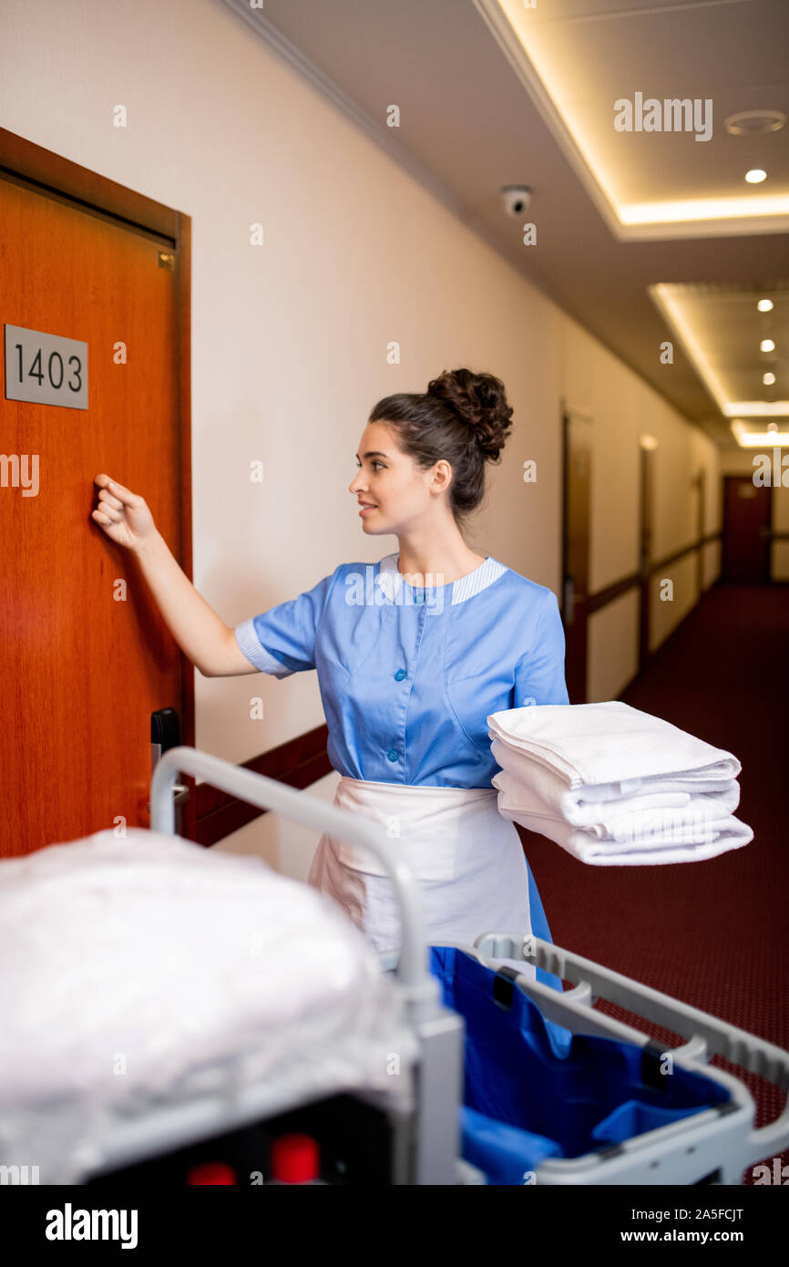 Pretty room maid in uniform holding stack of clean towels while knocking on wooden door in the morning Stock Photo