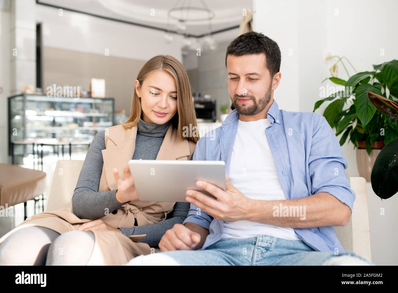 Casual young couple sitting on comfortable couch and scrolling in touchpad while relaxing in cozy cafe Stock Photo