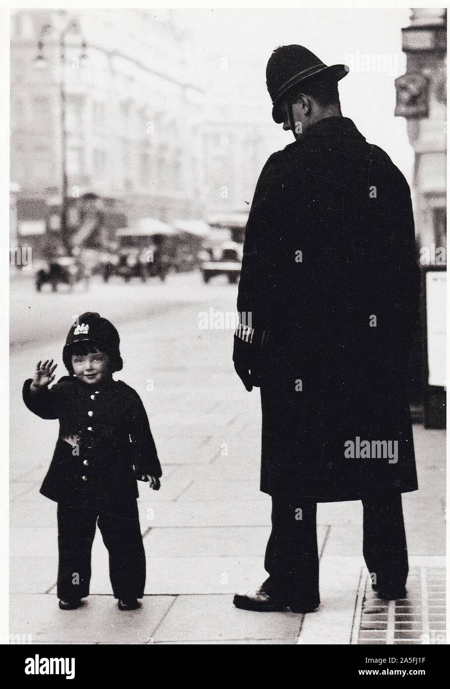 Vintage black and white postcard showing 1937 Bobbys (Policemen) with young boy dressed in uniform, view of 1930s London in background. Stock Photo