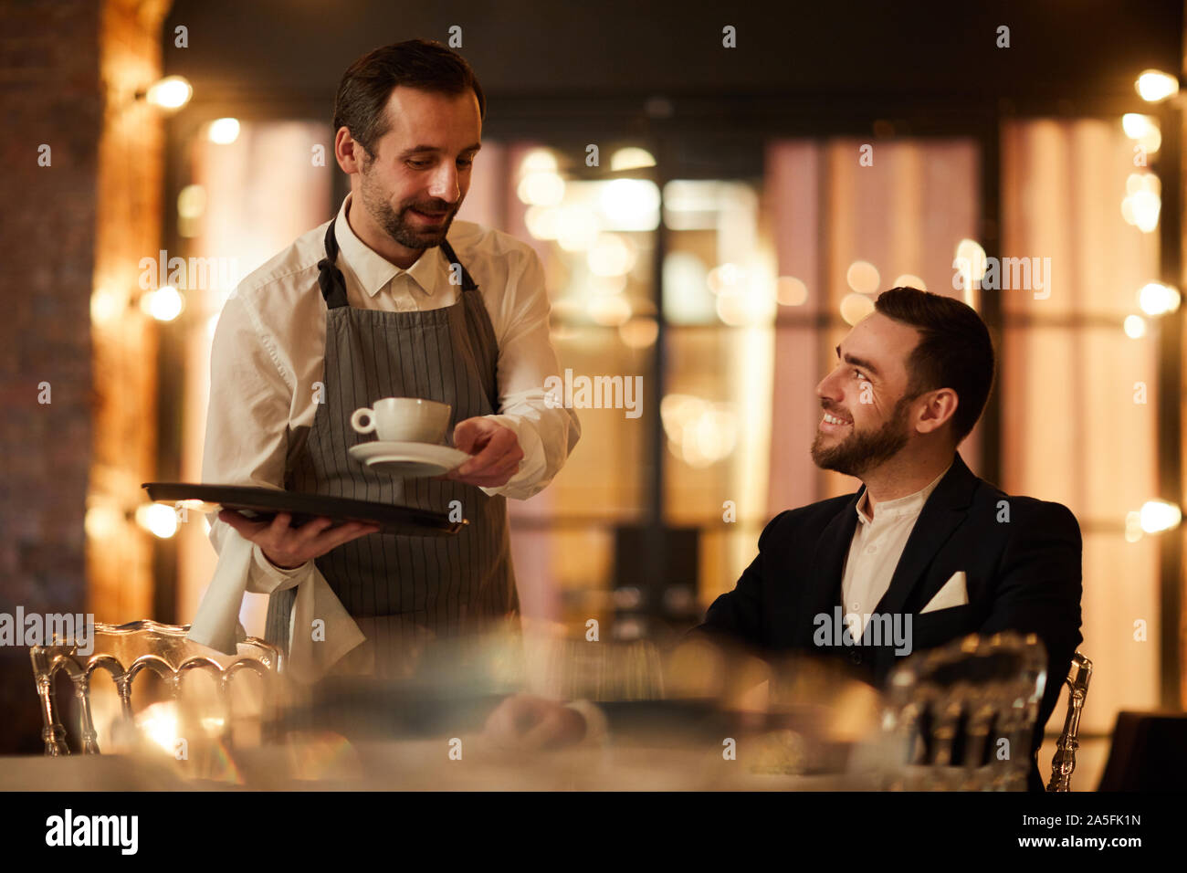 Side view portrait of handsome bearded businessman talking to waiter bringing coffee and smiling happily in luxury restaurant Stock Photo