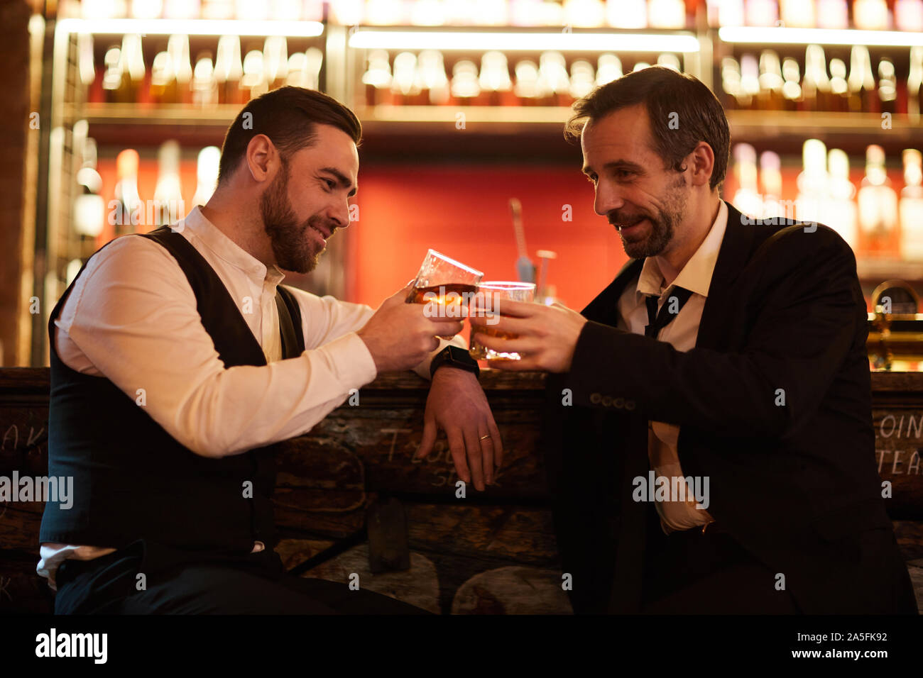 Side view portrait of two businessmen drinking whiskey and clinking glasses while sitting by bar counter and relaxing after work Stock Photo