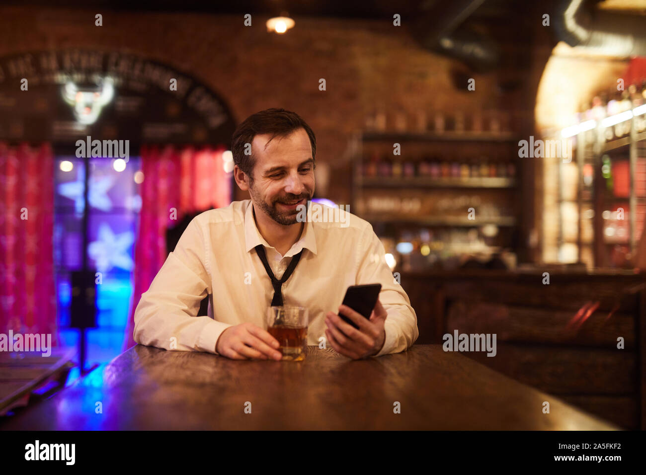 Portrait of mature businessman drinking alcohol in bar after work and smiling looking at smartphone screen, copy space Stock Photo