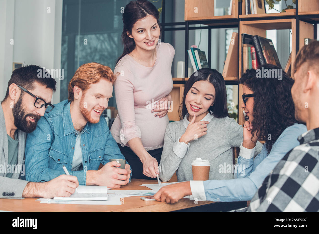 Startupers working together at office sitting looking at documents cheerful while pregnant woman standing pointing at idea for project Stock Photo