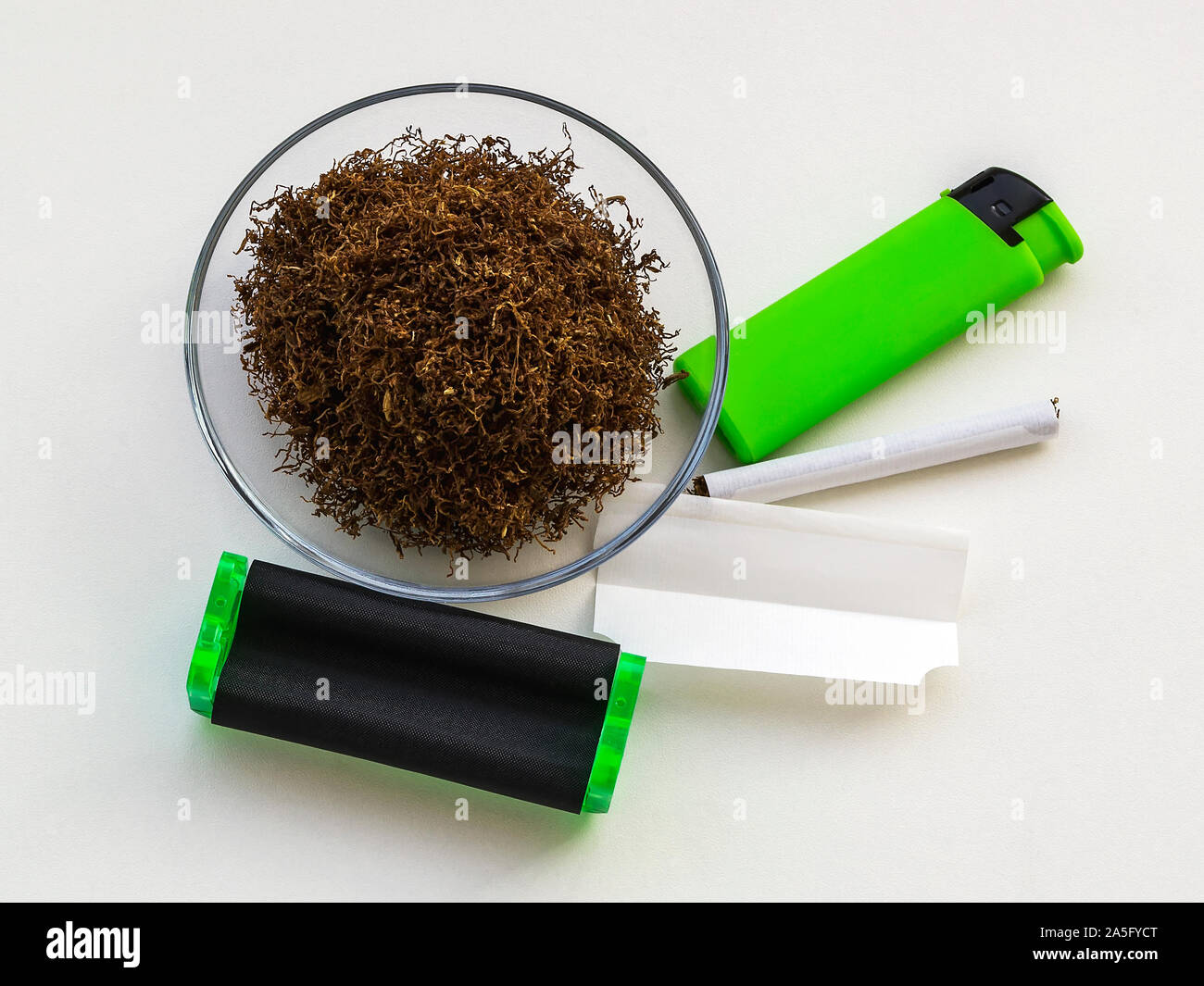 tobacco for making cigarettes Pile of tobacco in glass saucer, green cigarette rolling machine, paper,  one cigarette and green lighter on a white table. Making cigarettes Stock  Photo