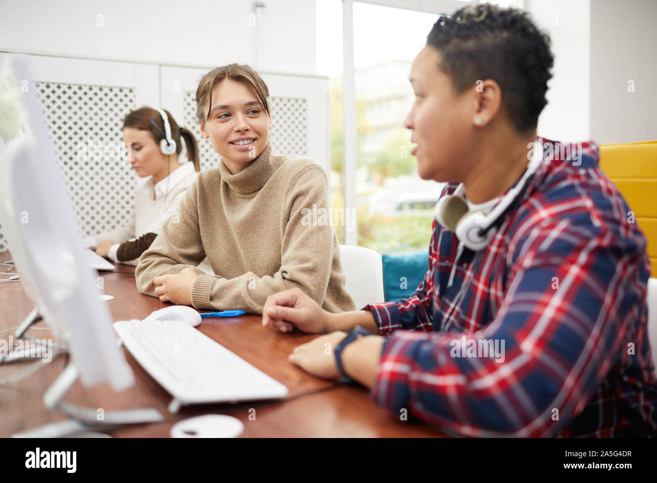 College students sitting in row and chatting while studying in computer class, focus on smiling young woman talking to friend, copy space Stock Photo