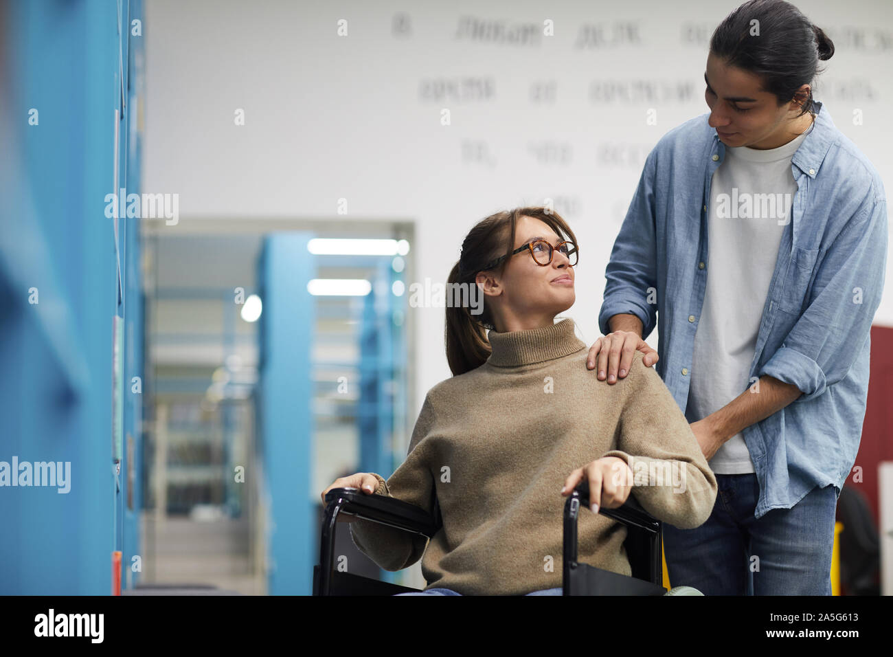 Portrait of young couple, woman in wheelchair, looking at each other and smiling while standing by shelves in library Stock Photo