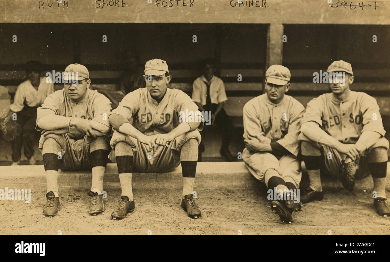 Photograph shows George Herman 'Babe' Ruth, Ernest G. 'Ernie' Shore, George 'Rube' Foster, and Dellos 'Del' Gainer, facing front, wearing Boston Red Sox baseball team uniforms, sitting on a low wall in front of a dugout. Stock Photo