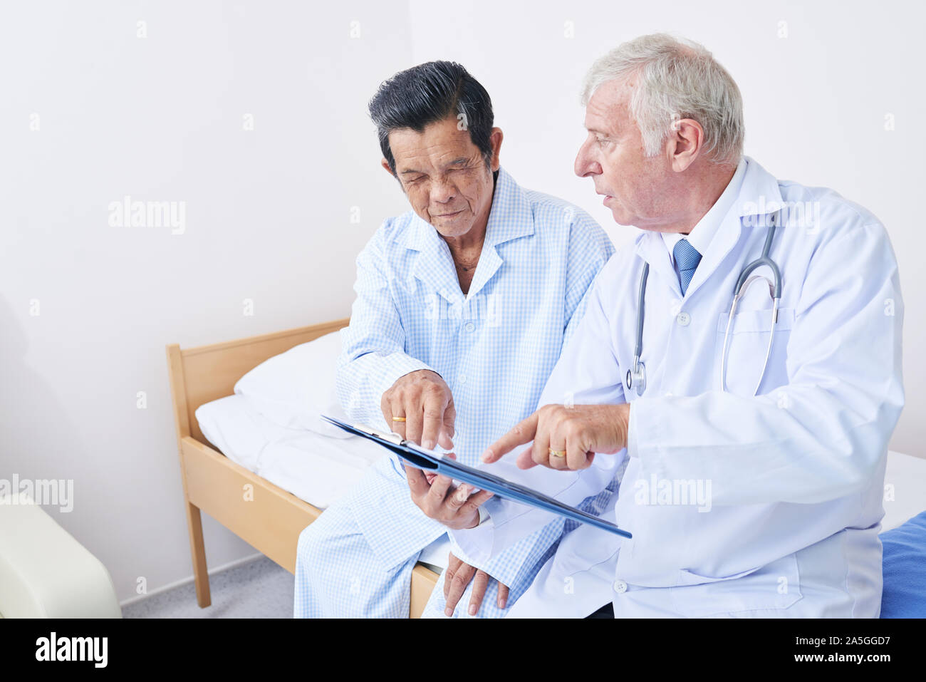Senior doctor sitting on bed of elderly Vietnamese patient and telling him diagnosis and prescribed regimen Stock Photo