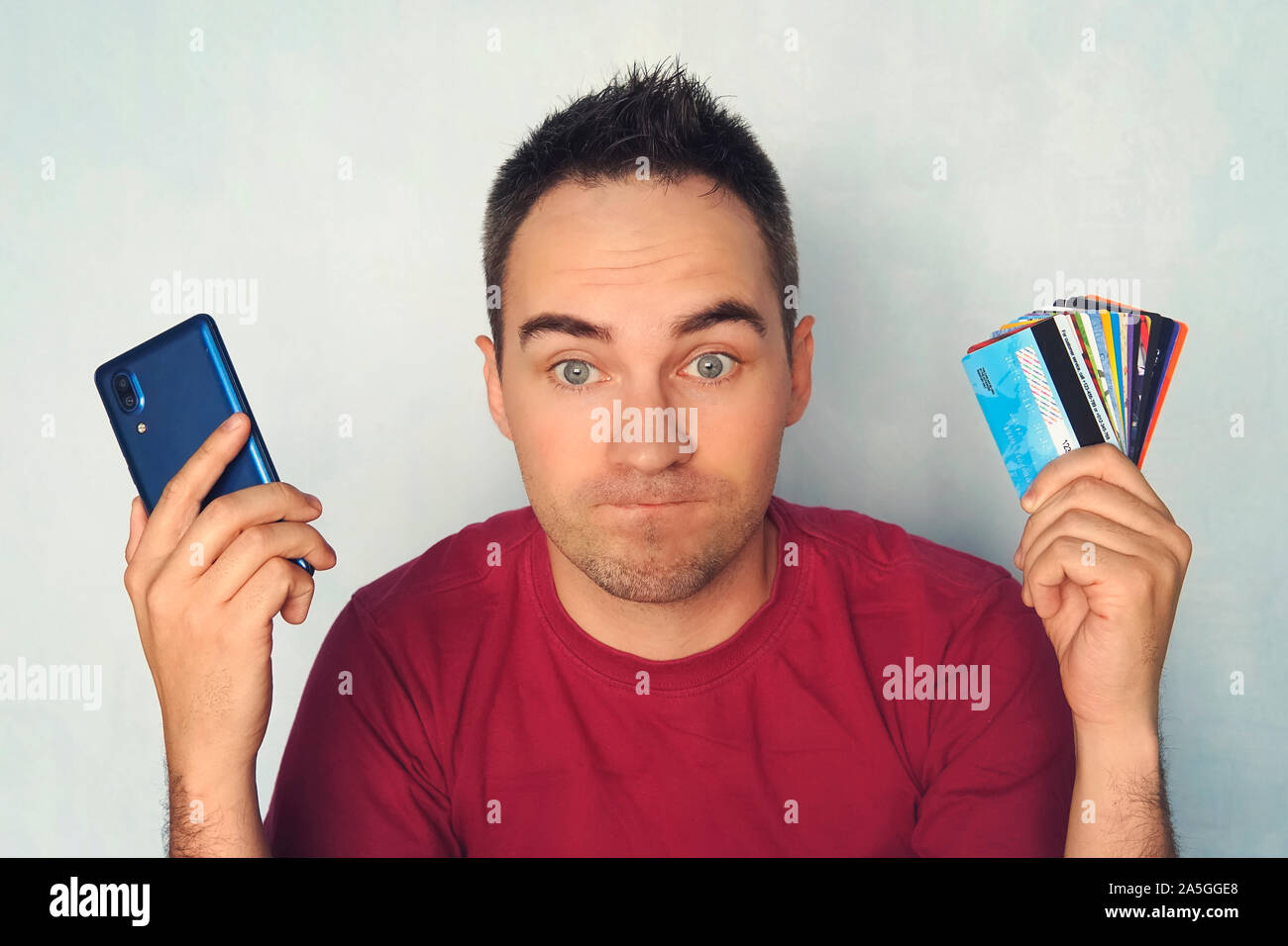 Guy Chooses A Credit Card To Pay Bewilderment On The Man S Face When Paying Through A Mobile Phone A Lot Of Plastic Cards In Hand Cell Phone For Ba Stock Photo