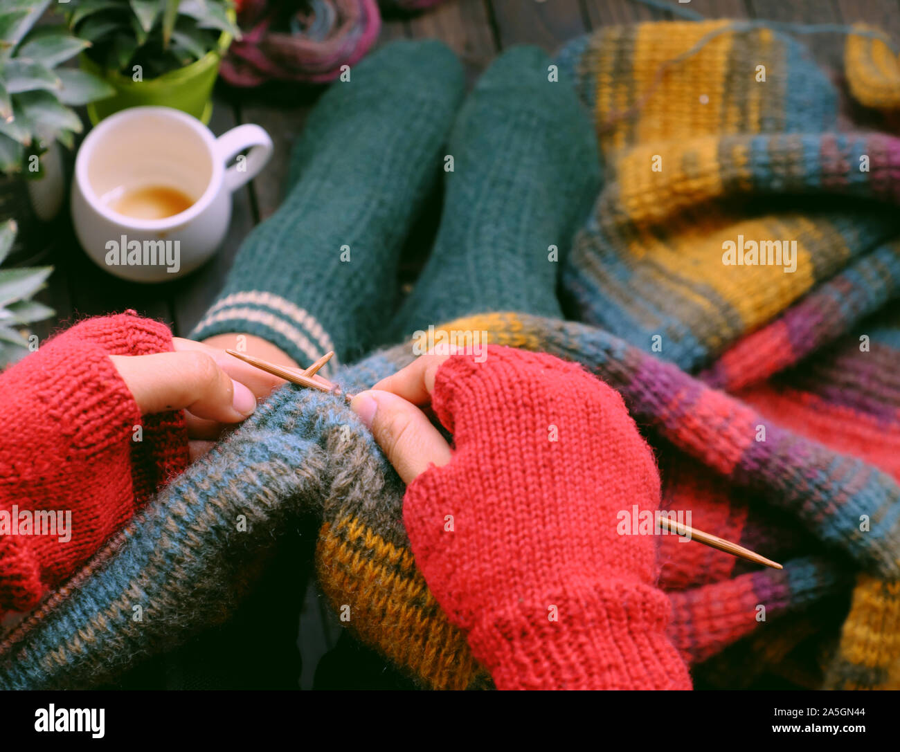 Top view woman feet in socks, sitting home balcony, hand with gloves hold knitting needle, knit colorful wool scarf, meaningful handmade winter gift Stock Photo