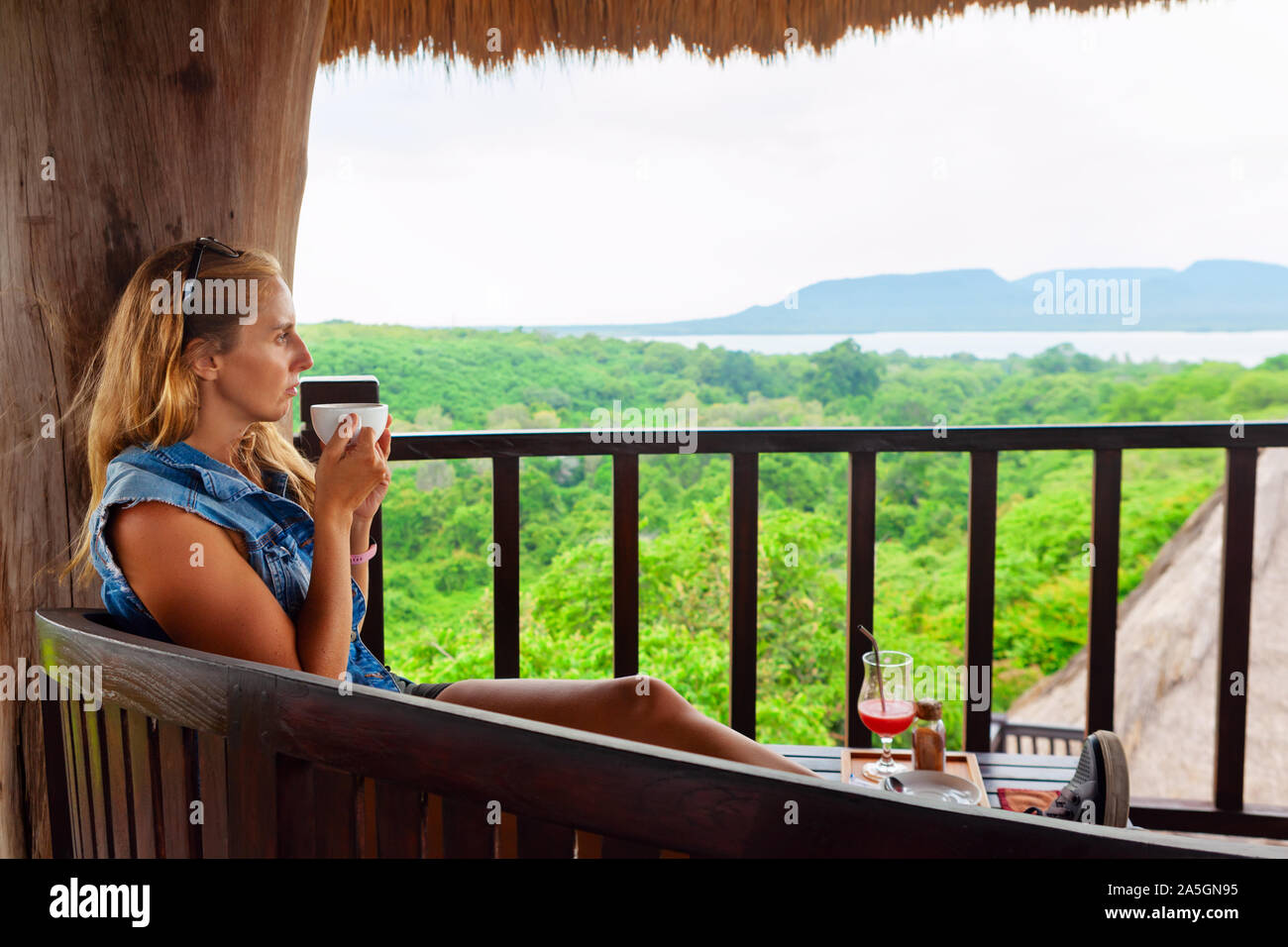 Happy family travel, explore rainforest near Pemuteran. Young woman drink morning coffee and relax on lounge veranda with tropical jungle view. Stock Photo