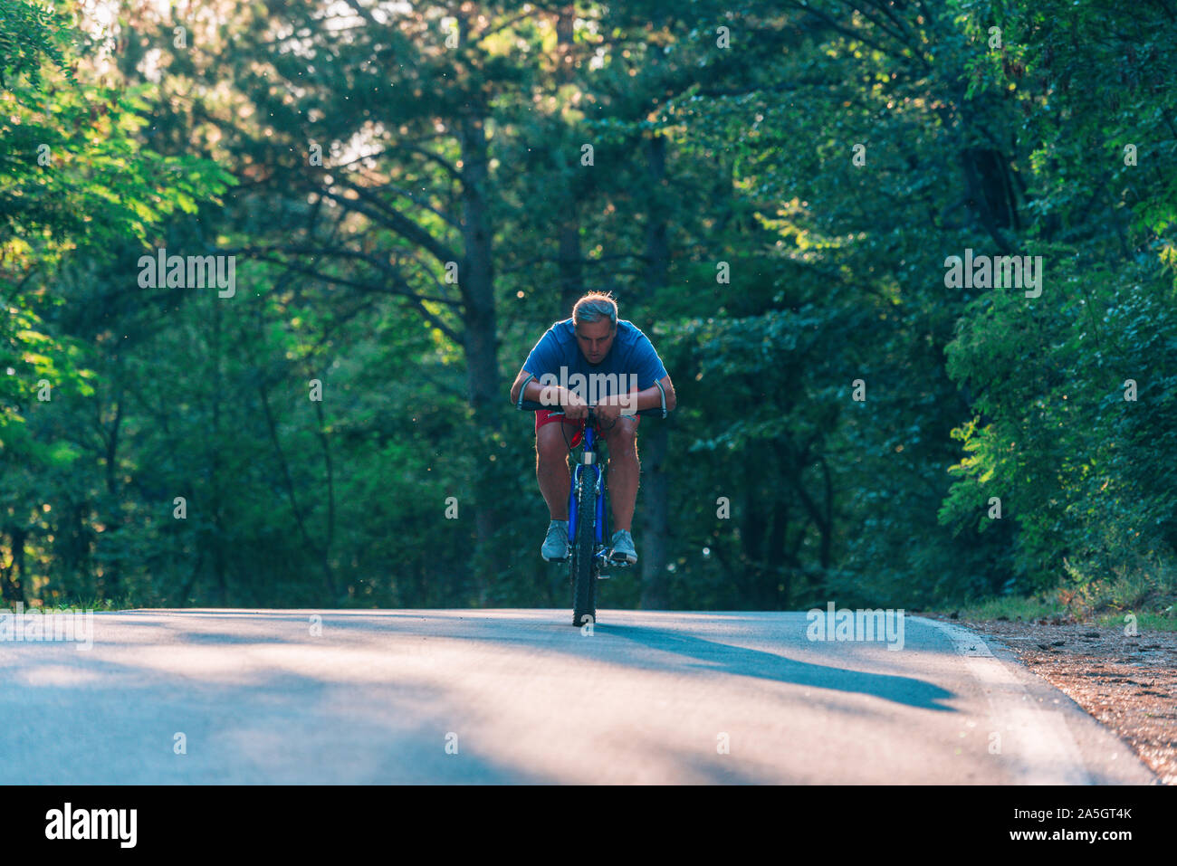 Older cyclist in his mid-40s riding a bike on a road through the woods ...
