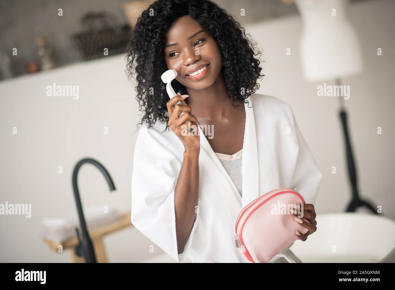 Dark-eyed woman smiling while using facial massage brush Stock Photo