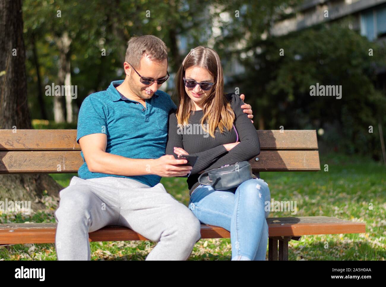Beautiful young couple sitting on the bench in park using mobile phone ...