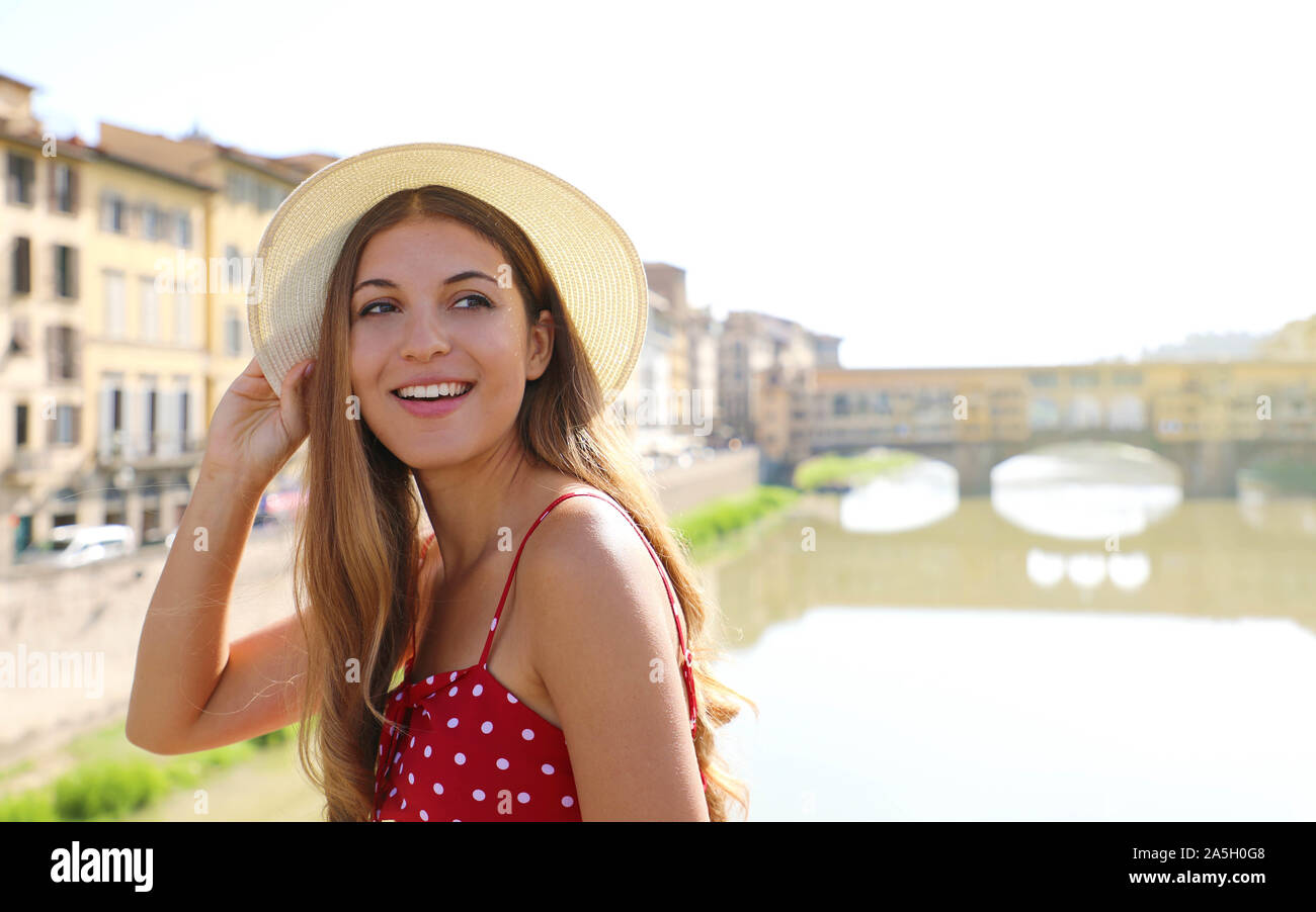 Beautiful smiling girl in Florence with Ponte Vecchio bridge and Arno river on the background. Portrait of young woman with hat and dress visiting Tus Stock Photo
