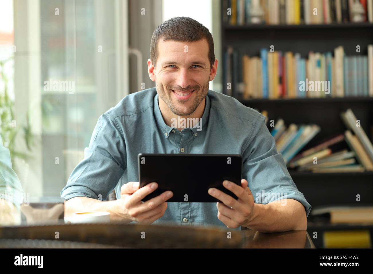 Front view portrait of a happy man holding tablet looking at camera sitting in a coffee shop or home Stock Photo